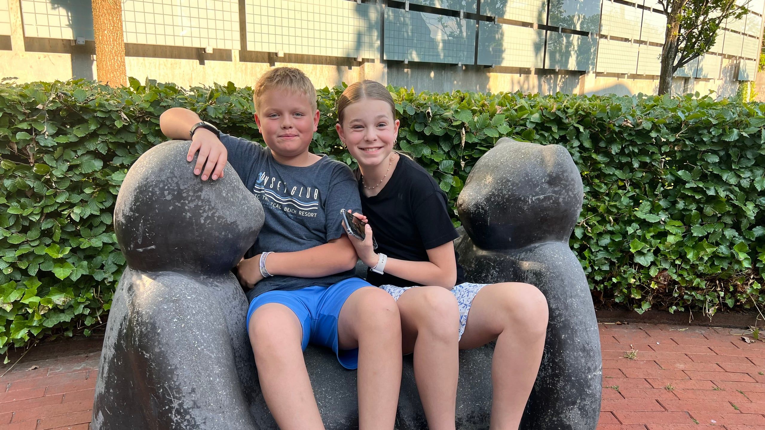 Noah og Naja på en skulpturbænk foran en hæk